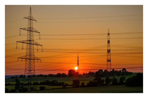 Foto im Querformat. Sonnenuntergang hinter einer grünen hügeligen Wiese mit vielen Bäumen und Büschen. Auf der Wiese stehen mehrere Hochspannungsmasten, die mit vielen Kabeln verbunden sind. Der Himmel ist goldgelb, weiter oben bläulich. Die Sonne ist knapp über dem Horizont, mittig. 