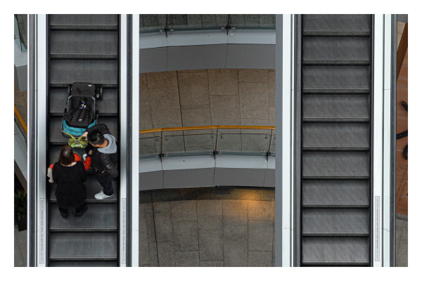 Foto im Querformat. Zwei Rolltreppen n einem Einkaufszentrum von oben fotografiert. Jeweils eine Rolltreppe links und rechts, dazwischen sind die verschiedenen Ebenen des Einkaufszentrums. Auf der linken Rolltreppe fährt eine Paar mit Kinderwagen nach oben. 