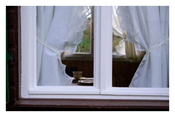 Foto im Querformat. Blick durch ein Fenster. Auf einem Tisch ist ein Kaffeegedeck. Am Fenster sind weiße Vorgänge. 