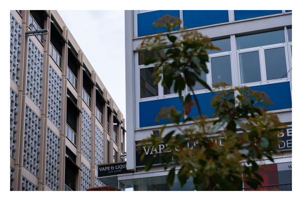 Foto im Querformat. Zwei Bürogebäude, eines links im Bild, mit vielen dicken Glaskacheln. Das andere rechts im Bild, mit blauer Verkleidung.  Die jeweiligen Kanten der Häuser treffen sich grob in der Mitte des Bildes. Vor dem blauen Gebäude ragen Äste mit Blättern ins Bild, die auf den ersten Blick palmenartig aussehen. Auf dem blauen Gebäude ist ein Schild auf dem Vape & Liquor steht. 