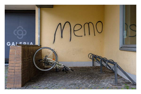 Foto im Querformat. Ein Fahrradständer in der Ecke eines Hauses. Rechts an der Wand steht der Ständer, gradeaus an der Wand ist das Wort Memo lieblos an die Wand gesprüht. Links ist eine kleine Mauer vor der ein Fahrrad seitlich auf dem Boden liegt. 