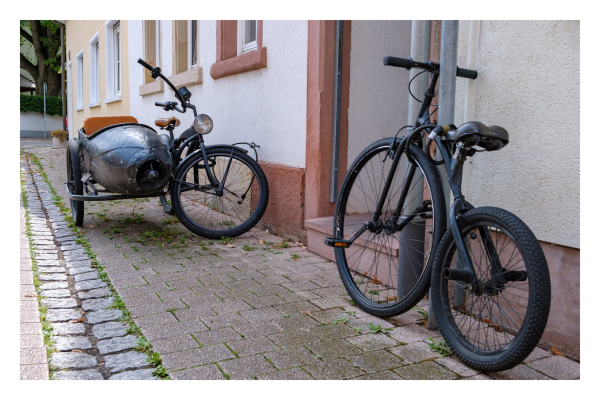 An einer Wand steht ein Hochrad, also ein Fahrrad, bei den der vordere Reifen wesentlich größer als der hintere ist und zudem die Pedale am großen Vorderrad sind. Allerdings ist dieses Hochrad wohl für Kinder ausgelegt, da es sehr klein ist. Ebenfalls an der Wand steht ein Fahrrad mit Beiwagen. 