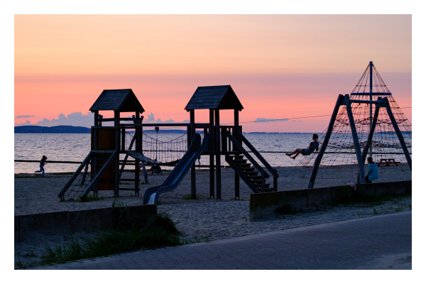 Foto im Querformat. Sonnenuntergang am Meer. Am Strand sind mehrere Spielgeräten wie eine Schaukel und ein Klettergerüst. Eine Person schaukelt, eine andere rennt über den Strand. Der Himmel ist in gelb und rot getaucht, der Spielplatz und die Personen zeichnen sich fast nur als Schatten ab. 