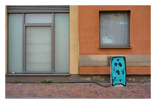 Foto im Querformat. Ein Gehweg vor zwei Hauswänden. Die eine Wand besteht nur aus einem großen Fenster, die andere ist verputzt mit einem kleinen Fenster. Unter diesem Fenster ist ein Stromkasten auf dem ein blaues gespenstartiges Wesen gemalt ist. Dieses hat erschrocken den Mund offen und schaut auf die linke untere Ecke des Kastens. An dieser Stelle kommt ein dickes Kabel aus dem Kasten, welches über den Gehweg und an den Häusern entlang läuft. 