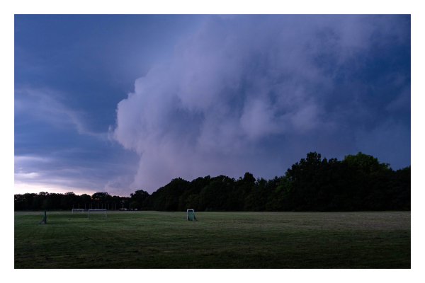 Foto im Querformat. Eine große Wiese, auf der mehrere Fußballtore stehen. Am Horizont ist Wald. Am Himmel sind riesige bedrohliche Wolken, die nach rechts hin immer dunkler werden. 