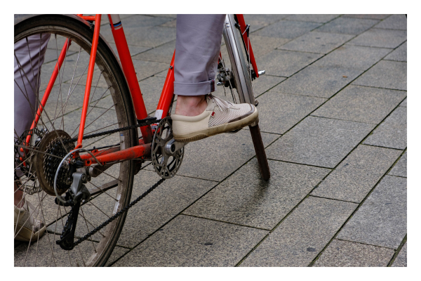 Foto im Querformat. Der untere Teil eines roten alten Fahrrads. Eine Person sitzt darauf und steht mit einem Fuß am Boden. Der andere Fuß steht auf dem Pedal. Die Person hat eine hochgekrempelte Hose an, keine Socken und Segelschuhe. 