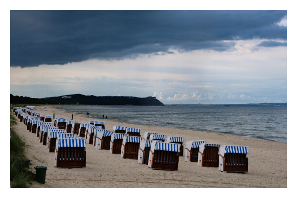 Foto im Querformat. Ein Strand zieht sich bis zum Horizont, an dem eine dunkle Küste ins Meer ragt. Am Strand sind Dutzende Strandkörbe, alle aus Holz, braun, mit blau weiß gestreiftem Dach. Drei Reihen ziehen sich den Strand entlang. Das Wetter ist bedeckt mit dunklen Wolken, die Strandkörbe sind leer. 