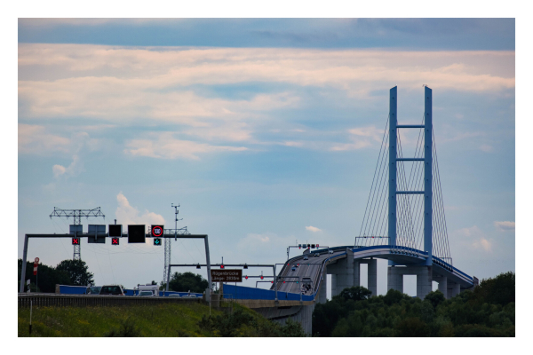 Foto im Querformat. Blick auf die Rügenbrücke, die von Festland auf die Insel Rügen führt. Die Brücke verläuft in einem Bogen, am höchsten Punkt sind links und rechts zwei hohe Säulen, an denen Stahlseile befestigt sind, die die Brücke tragen. 
Der Himmel ist bewölkt. 