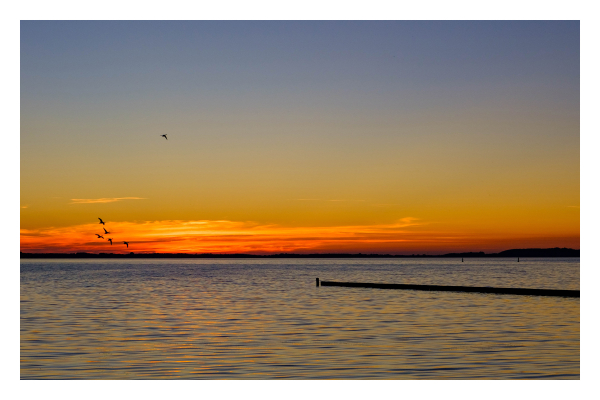 Foto im Querformat. Blick aufs Meer mit Sonnenuntergang. Die Sonne ist gerade hinter dem Horizont verschwunden, beleuchtet die Wolken am Horizont aber noch orange-rot. Vor den Wolken sind die Silhouetten einiger Vögel auszumachen. Von rechts ragt ein dünner Steg ins Bild und geht fast bis zur Mitte. 