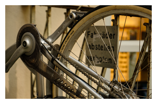 Foto im Querformat. Detailaufnahme eines Fahrrads, welches auf dem Kopf steht. Im. Und sind die Pedale, die Kette und ca. ein Drittel des Hinterrades. Durch die Speichen ist ein Schild an einem Geländer zu sehen. Auf diesem steht Fahrräder abstellen verboten. 