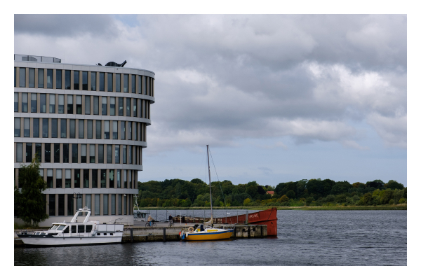 Foto im Querformat. Ein Bürogebäude, welches nach oben hin breiter wird. Davor ist ein Steg, im Wasser liegen drei Boote. Links ein Motorboot, rechts ein gelbes kleines Segelboot und ein rotes größeres Segelschiff. Bis auf die zwei Segelboote ist das restliche Bild in Grautönen gehalten. Am gegenüberliegenden Ufer sind Wälder. Der Himmel ist bewölkt. 