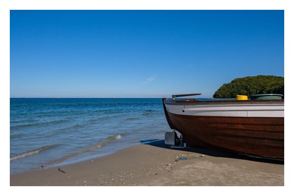 Foto im Querformat. Strand mit Blick aufs Meer. Rechts im Bild ist die vordere Hälfte eines Bootes, der Rumpf ist braun mit oben einem weißen Streifen. Die Spitze des Bootes geht genau bis zum Wasser. Der Horizont teilt das Bild genau in der Mitte, der Himmel ist strahlend blau. 