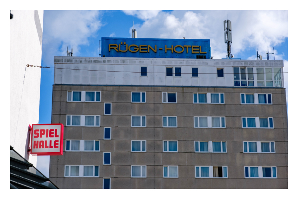 Foto im Querformat. Die obere Hälfte des Rügen-Hotels. Ein rechteckiger dunkelgrauer Betonklotz mit vielen rechteckigen Fenstern. Auf dem Dach ist ein blaues Schild auf dem Rügen-Hotel steht. Daneben ein Sendemast. Im Vordergrund ist an einem anderen Gebäude ein rot-weißes Schild auf dem Spielhalle steht. Der Himmel über dem Hotel ist leicht bewölkt. 