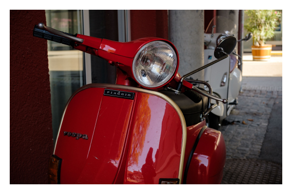 Foto im Querformat. Vorderansicht einer knallroten Vespa, ein Motorroller der Marke Piaggio. Die Vespa steht an einer Hauswand, weiter hinten steht eine weitere weiße Vespa. Im Lack des Motorrollers spiegele ich mich, gebückt beim fotografieren. 