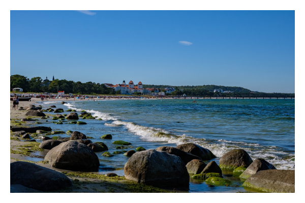 Foto im Querformat. Ein lang gezogener Strand, der einen Bogen nach rechts verläuft. Am Horizont ist der Strandprt Binz. Ein langer Fußgängersteg führt aufs Meer. Im Vordergrund sind einige Felsen, weitere sind weiter vorne zwischen Wasser und Strand. Der Strand ist voll mit Menschen, einige sind auch im Wasser. Weiße Gischt kommt gerade am Strand an und zieht sich parallel dazu bis zum Horizont. Der Himmel ist bis auf zwei kleine Wolken strahlend blau. 