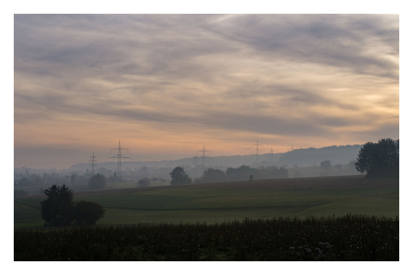 Foto im Querformat. Eine Landschaft in Nebel getaucht. Wiesen, Bäume, Wälder, Strommasten. Alles ist nur als Kontur zu erkennen. Der Himmel ist mit Wolken bedeckt, die von der aufgehenden Sonne gelblich angestrahlt werden. Die Sonne ist außerhalb des Bildes. 