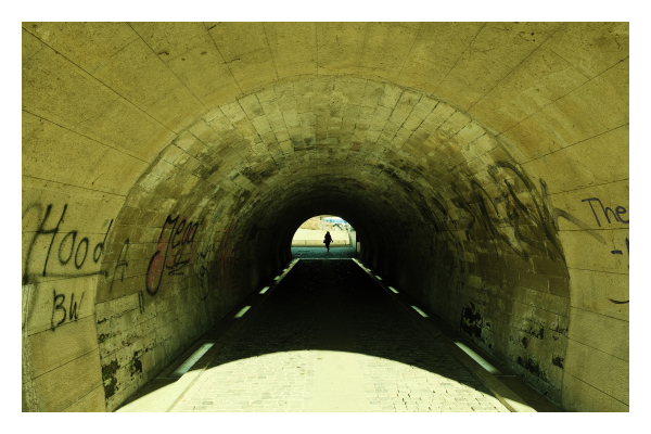 Foto im Querformat. Blick in einen Fußgängertunnel. Am anderen Ende läuft mittig eine einzelne Person. Im Tunnel ist Schatten, links und rechts sind helle Streifen am Boden. Vor dem Tunnel fällt in emblem Halbkreis Licht auf den Boden. Die Wände des Tunnels sind vollgekritzelt mit schlechten Graffiti. 