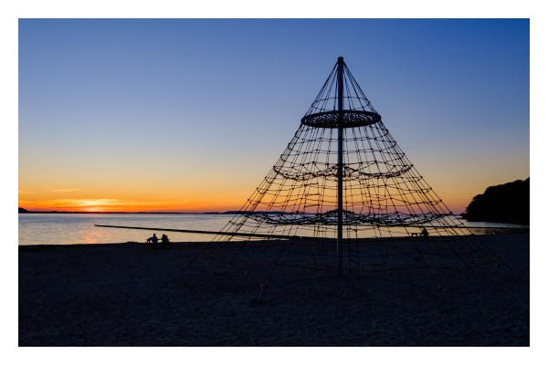 Foto im Querformat. Sonnenuntergang am Strand. Die Sonne ist am Horizont nur noch zu erahnen, sie taucht den Himmel in gelb-rotes Licht. Der Strand ist nur als Schatten zu erkennen, zwei Personen sitzen im Sand und schauen sich den Sonnenuntergang an. Im Vordergrund ist noch die Silhouette eines kegelförmigen Klettergerüst für Kinder. 