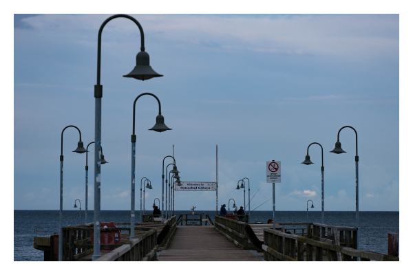 Foto im Querformat. Blick aufs Ende eines Besucherstegs an der Ostsee. Links und rechts sind Straßenlaternen, die oben gebogen sind. Ebenfalls links und rechts stehen Menschen die im Meer angeln. Am Ende des Stegs ist über den Steg ein Schild, auf dem „Willkommen im Ostseebad Göhren“ steht. Der Himmel ist bedeckt und sehr düster. 