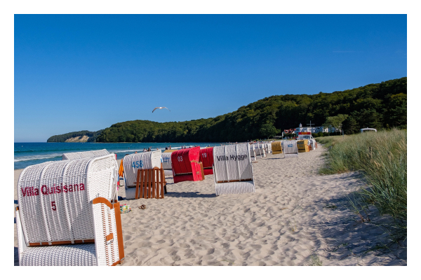 Foto im Querformat. Ein Strand auf dem viele Strandkörbe stehen. Überwiegend weiße, ein paar sind rot. Rechts ist Gras der Dünen, links hinter den Strandkörben ist das blaue Meer. Am Horizont sind mit Bäumen bewachsene Klippen. Über den Strand fliegt eine einzelne Möwe Richtung Meer. 