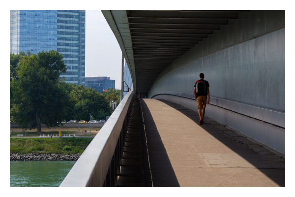 Foto im Querformat. Eine Brücke über einen Fluss in Blickrichtung der Brücke. Es ist eine überdachte Fußgängerbrücke, die den rechten Teil des Bildes ausmacht. Führende Linien Richtung Horizont sind das Geländer, das Dach und jeweils der Schatten, der von beidem auf den Fußweg geworfen wird. Zwischen den Schatten ist ein breiter Streifen, der von der Sonne beschienen wird. Eine Person läuft über die Brücke, auf dem engen Streifen Schatten, welcher das Dach wirft. Links im Bild ist der Fluss, Bäume und ein großes Bürogebäude. 