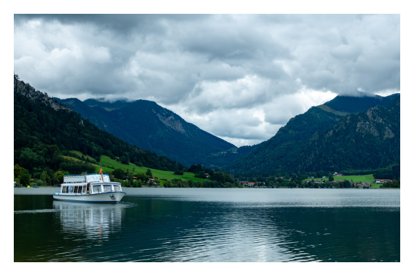 Foto im Querformat. Ein See, darauf fährt eine kleine Fähre von links nach rechts. Hinter dem See erstreckt sich ein Tal, links und rechts hohe Berge. Die Berge werfen ihren Schatten auf den See, in der Mitte kommt die Sonne durch und erhellt das Wasser. Der Himmel ist mit Wolken verhangen. 