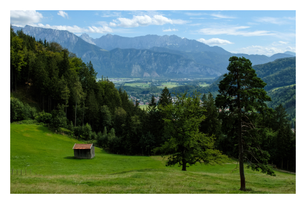 Foto im Querformat. Blick von einem Berg übers Alpenvorland auf die Alpen in Österreich. Um Vordergrund ist eine Bergwiese mit einem kleinen Haus und zwei einzelnen Bäumen. Dahinter ist Wald, wiederum dahinter der Blick ins Tal. Es ist Sommer, der Himmel ist leicht bewölkt. 