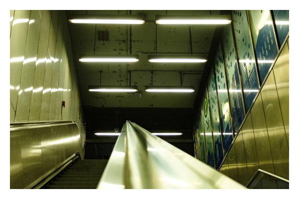 Foto im Querformat. Blick von unten über das Geländer einer Rolltreppe. Das Geländer ist mittig im Bild, links die Rolltreppe, rechts die normale Treppe. An der Decke sind mehrere Lampen mit Neonröhren, die alles in ein grünliches harsches Licht einbetten. Die Lampen Spiegeln sind an den Wänden und im Geländer der Rolltreppe. 