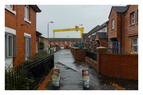 Foto im Querformat. Blick in ein Wohngebiet in Belfast. Alle Häuser sind aus rotbraunem Backstein. Der Blick der Kamera folgt einem Gehweg durchs Wohngebiet, vorne sind zwei Steinpfosten, die ein reinfahren mit dem Auto verhindern. Über den Häusern am Ende des Weges ist ein riesiger gelber Containerkran. Der Himmel ist bedeckt. 