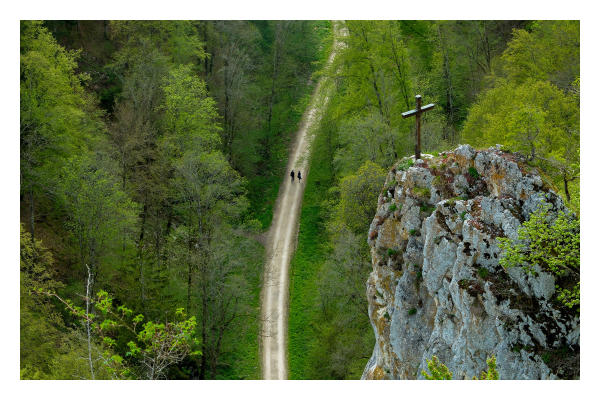 Foto im Querformat. Blick in ein Tal von oben. Rechts im Bild, am Berg, ragt ein großer Fels ins Bild. Auf seiner Spitze ist ein Gipfelkreuz. Mittig im Bild verläuft unten im Tal ein geschwungener Kiesweg. Darauf laufen zwei Personen, die von oben sehr klein aussehen. Links und rechts des Weges ist grüner Wald. 