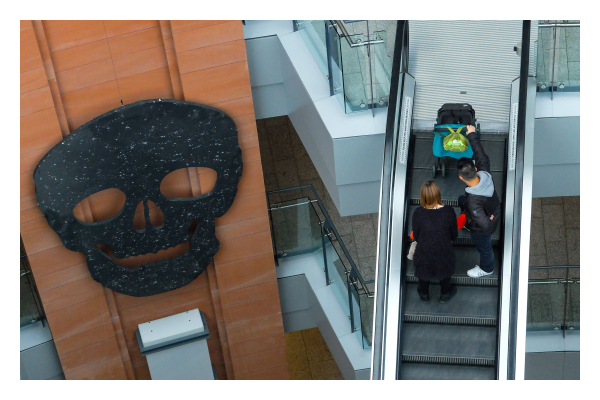 Foto im Querformat. Blick von oben in eine Shoppingmall. Rechts ist eine Rolltreppe auf der zwei Menschen mit einem Kinderwagen sind. Links ist rotbraune Wand an der ein riesiger schwarzer Totenkopf hängt. Er ist so groß wie ein Stockwerk der Mall. 