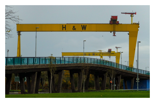 Foto im Querformat. Eine große geschwungene Brücke geht von links nach rechts durchs Bild. Anfang und Ende sind jeweils nicht zu sehen. Die Brücke hat ein grünes Geländer auf beiden Seiten. Über der Brücke erheben sich zwei riesige gelbe Containerkräne auf denen H & W steht. Der Himmel ist bedeckt. 