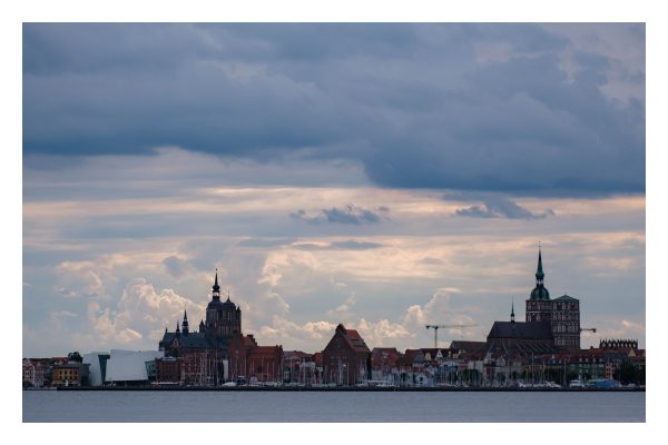 Foto im Querformat. Der Hafen Stralsunds von der anderen Seite des Wassers aus fotografiert. Viele rote Backsteinbauten, Kirchtürme, Kräne, Segelschiffe. Links, groß in weiß das Ozeaneum, ein modernes Gebäude. Der Himmel ist bewölkt. 