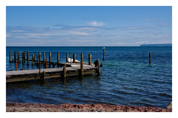 Foto im Querformat. Küste, ein dünner Streifen Strand unten im Bild, dahinter leicht gekräuseltes dunkelblaues Meer. Von links ragt ein hölzerner Steg ins Bild, der wiederum links abbiegt. An diesem sind viele Pfosten, um ein Boot daran anzubinden. Ein einziges Boot ist am Steg befestigt. Im Meer sind auch ein paar Pfosten, auf einem sitzt eine weiße Möwe. Der Himmel ist leicht bewölkt. 