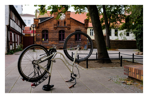 Foto im Querformat. Ein öffentlicher Platz in einer Stadt umringt von alten Gebäuden, teils Fachwerk, teils aus rotbraunen Backsteinen. Auf dem Platz steht ein Fahrrad mit tiefem Einstieg auf dem Kopf. Durch das vordere Rad, sieht man weitere Räder, die weiter hinten an einem Fahrradständer stehen. 