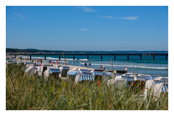 Foto im Querformat. Blick über einen Strand voller Strandkörbe. Im Vordergrund ist hohes Gras auf einer Düne, dann der Strand, schließlich das Meer. Vom Strand geht ein riesiger Steg ins Meer, dessen Ende nicht zu sehen ist. Der Himmel ist blau und wolkenlos. 