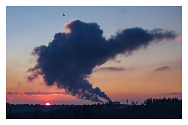 Foto im Querformat. Sonnenuntergang, der untere Teil des Bildes ist Wald, der im Schatten liegt. Darüber geht die Sonne unter, sie ist nur noch halb zu sehen. Der blau-rote Himmel wird von einer riesigen Rauchwolke überdeckt, die aus Schornsteinen kommt, die hinter dem Horizont sind. Der restliche Himmel ist wolkenlos. Ganz oben im Bild ist ein Flugzeug, welches auf die Rauchwolke zufliegt. 