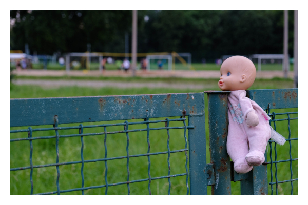 Foto im Querformat. An einem grünen Zaun aus Metall hängt eine Babypuppe. Diese wurde mit dem Hals zwischen zwei Pfosten geklemmt. Im Hintergrund ist Wiese auf der in der Unschärfe mehrere Menschen beim Fußball spielen zu erkennen sind. Es sieht ein bisschen so auf, als ob die Puppe zuschaut. 