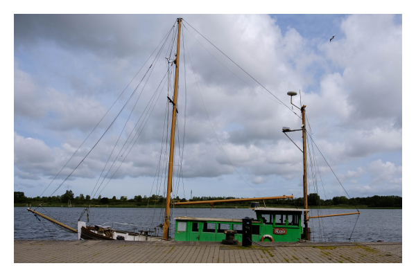 Foto im Querformat. Ein Segelboot liegt am Pier. Der untere Teil des Bootes ist unterhalb des Piers, es hat zwei Masten und eine grüne Kajüte. Die Segel sind eingefahren. Dahinter ist Wasser, man sieht das gegenüberliegende Ufer. Der Himmel ist bewölkt. 