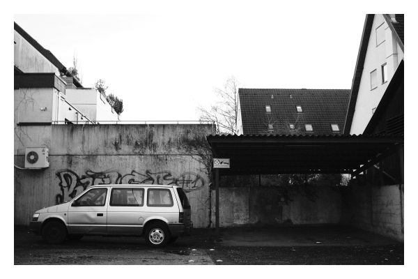 Foto im Querformat. Schwarzweiß Bild. Ein Hinterhof. Vor einer besprühten Mauer steht ein heller Kleinwagen. An der Mauer hängt eine Klimaanlage. Rechts daneben ist ein Vordach an der Mauer. Dieses wird von Pfosten gehalten. An einem Pfosten ist ein Parkverbotsschild mit dem Hinweis, dass abgeschleppt wird. Hinter der Mauer sind Dächer von Wohnhäusern zu sehen und der Himmel, der weiß ist. 