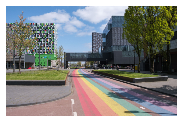 Foto im Querformat. Das Gelände der Universität in Utrecht/Niederlande. Von vorne nach hinten verläuft ein breiter zweispuriger Fahrradweg. Dieser ist gestreift wie ein Regenbogen. Links und rechts sind die hohen Gebäude der Universität, diese sind teilweise auch bunt kariert. 