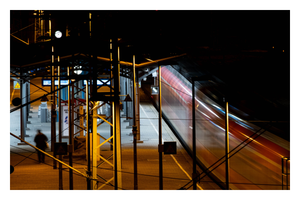 Foto im Querformat. Zoomaufnahme eines Gleises eines Bahnhofs. Es ist Nacht und dunkel, auf dem Bahnsteig sind viele Pfosten, Lampen und Säulen. Ein Zug fährt gerade los, durch die lange Belichtung hat dieser Bewegungsunschärfe. Links auf dem Bahnsteig ist eine einzelne Person, diese ist ebenfalls unscharf. 