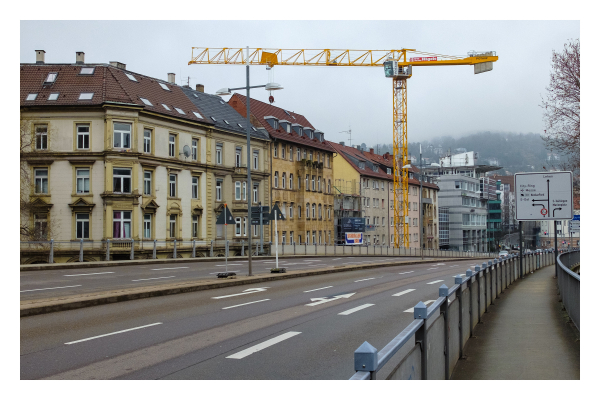 Foto im Querformat. Eine leere mehrspurige Hauptstraße in einer Großstadt. Sie verläuft von links unten nach rechts im Bild. Hinter der Straße sind mehrere Altbauten und ein großer gelber Kran. Weiter hinten erhebt sich ein Berg, der in Nebel getauft ist. Rechts neben der Straße verläuft ein Fußweg, der mit einem Geländer von der Straße abgetrennt ist. 