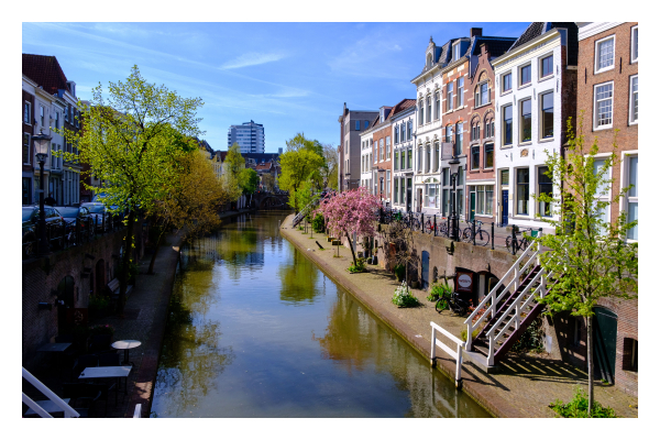 Foto im Querformat. Blick entlang einer Gracht in Utrecht. Mittig verläuft der Kanal, links und rechts sind kaum höher als der Wasserspiegel Ebenen. Auf diesen stehen Bäume und es gibt Türen. Über Treppen kommt man hoch auf die Straße, an diesen stehen viele Fahrräder. An den Straßen sind alte Gebäude, die alle unterschiedlich aussehen. Am Horizont ist ein einzelnes modernes Hochhaus. Alles ist menschenleer. Der Himmel ist leicht bewölkt. 