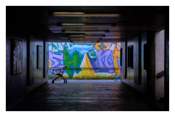 Foto im Querformat. Blick durch eine Unterführung. Es ist dunkel, an der Decke sind hintereinander Neonröhren, die kaum Licht geben. Rechts geht ein Treppenaufgang hoch, durch den etwas Licht fällt. Am Ende der Unterführung steht links ein einzelner fast leerer Einkaufswagen. Das Ende der Unterführung wird von der Sonne beleuchtet, dort ist eine Wand die voll mit Graffiti ist. 