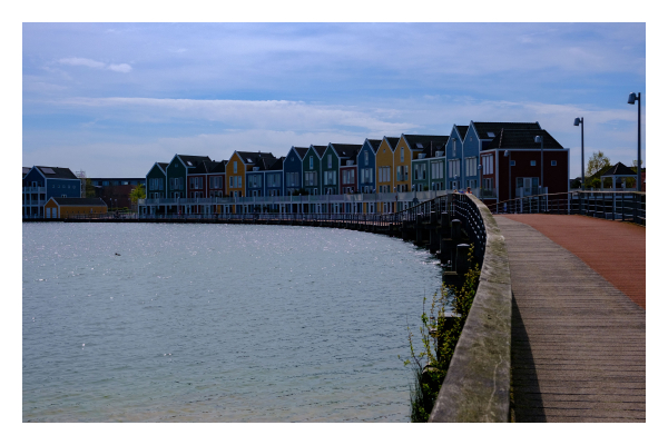 Foto im Querformat. Blick einen Steg entlang. Der Blick der Kamera folgt dem hölzernen Geländer welches einen leichten Bogen nach links macht. Auf der rechten Seite des Steges  sind mehre Wohnhäuser in unterschiedlichen Farben. Blau, gelb und rot. Links vom Steg ist Wasser. 