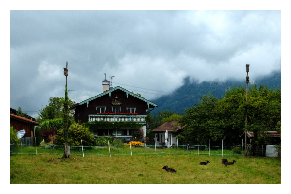 Foto im Querformat. Ein bayerisches Bauernhaus, vertäfelt mit Holz. Davor ein Garten, daneben ein kleineres Haus. Vor dem Garten liegen ein paar Ziegen im Gras. Hinter dem Haus lässt sich ein bewaldeter Berg erahnen, dieser ist aber komplett von Wolken bedeckt. 