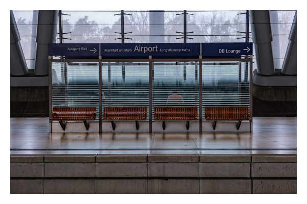 Foto im Querformat. Wartebänke an einem Bahngleis frontal fotografiert. Vier Bänke, dahinter eine Wand aus milchigem Plexiglas, dahinter nochmal vier Bänke. Auf einer der Bänke hinter der Wand ist eine einzelne Person auszumachen. Diese sitzt und schaut nach unten. Alles im Foto ist sehr symmetrisch, auch die Balken an den Fenstern im Hintergrund. 