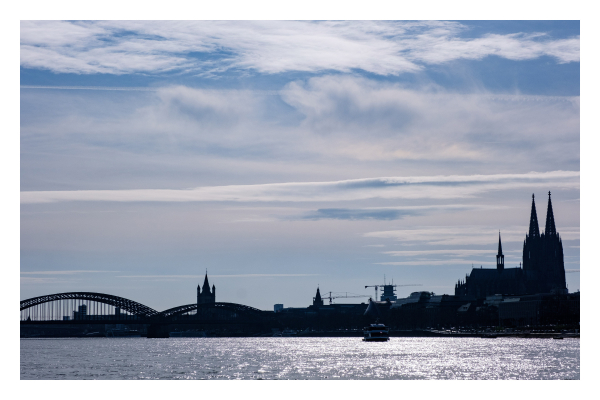 Foto im Querformat. Die Skyline von Köln über den Rhein fotografiert. Rechts ist der Dom, ganz links eine Bogenbrücke. Auf dem Rhein fährt ein Schiff. Die ganze Skyline ist im Schatten und quasi schwarz. Der Himmel ist leicht bewölkt und auf dem Wasser spiegeln sich die Sonnenstrahlen. 