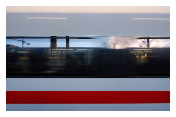 Foto im Querformat. Ausschnitt eines ICE Zuges, seitlich. Der Zug ist weiß, unterhalb des Fensters ist ein roter Streifen. Im Fenster des vorbeifahrenden Zuges spiegelt sich ein Bahnhof, im Hintergrund ist ein Kran. Bei genauerem hinschauen sieht man auf einem Schild Stuttgart stehen. Das weiß und rot des Zuges und die dunklen Scheiben ergeben ein symmetrisches Streifenmuster. 